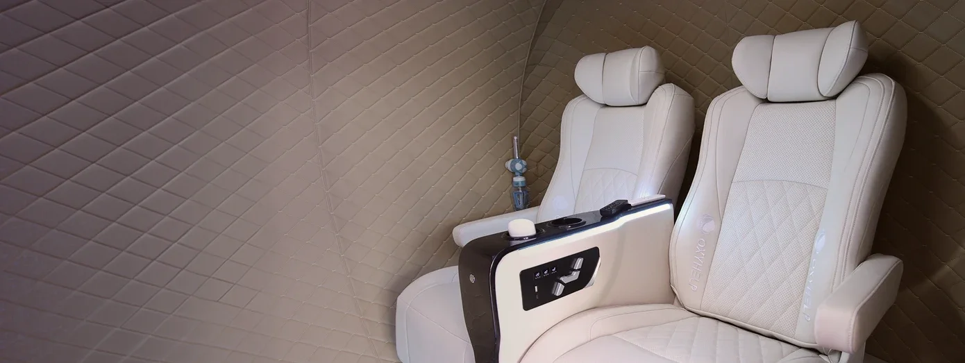 Two cream recliner seats inside an OxyLife chamber, with the armrest control panel