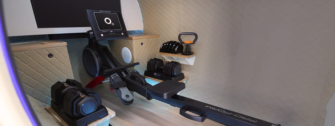 Rowing machine, adjustable dumbbells and a kettlebell inside an OxyLife Gym chamber, with a screen above